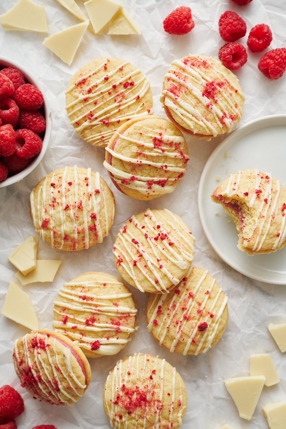 several Raspberry Cream Sandwich Cookies on a white background, next to a bowl of fresh raspberries. 