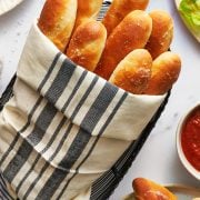 copycat olive garden breadsticks recipe wrapped in a linen towel in a bread basket with marinara sauce on the side.