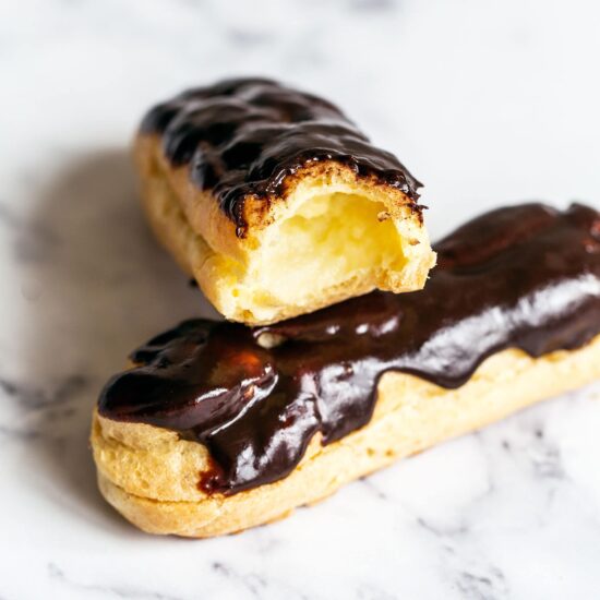 Two chocolate eclairs on marble, with a bite taken out of one to show pastry cream inside