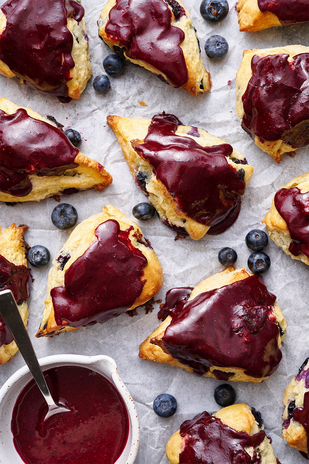 glazed blueberry scones on parchment paper, with fresh blueberries scattered throughout