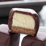 three chocolate-covered cheesecake bites stacked, with one cut in half so you can see the cheesecake and graham cracker crust base inside.