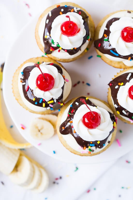 Banana cupcakes with vanilla buttercream frosting, chocolate ganache, sprinkles, whipped cream, and a cherry on top.