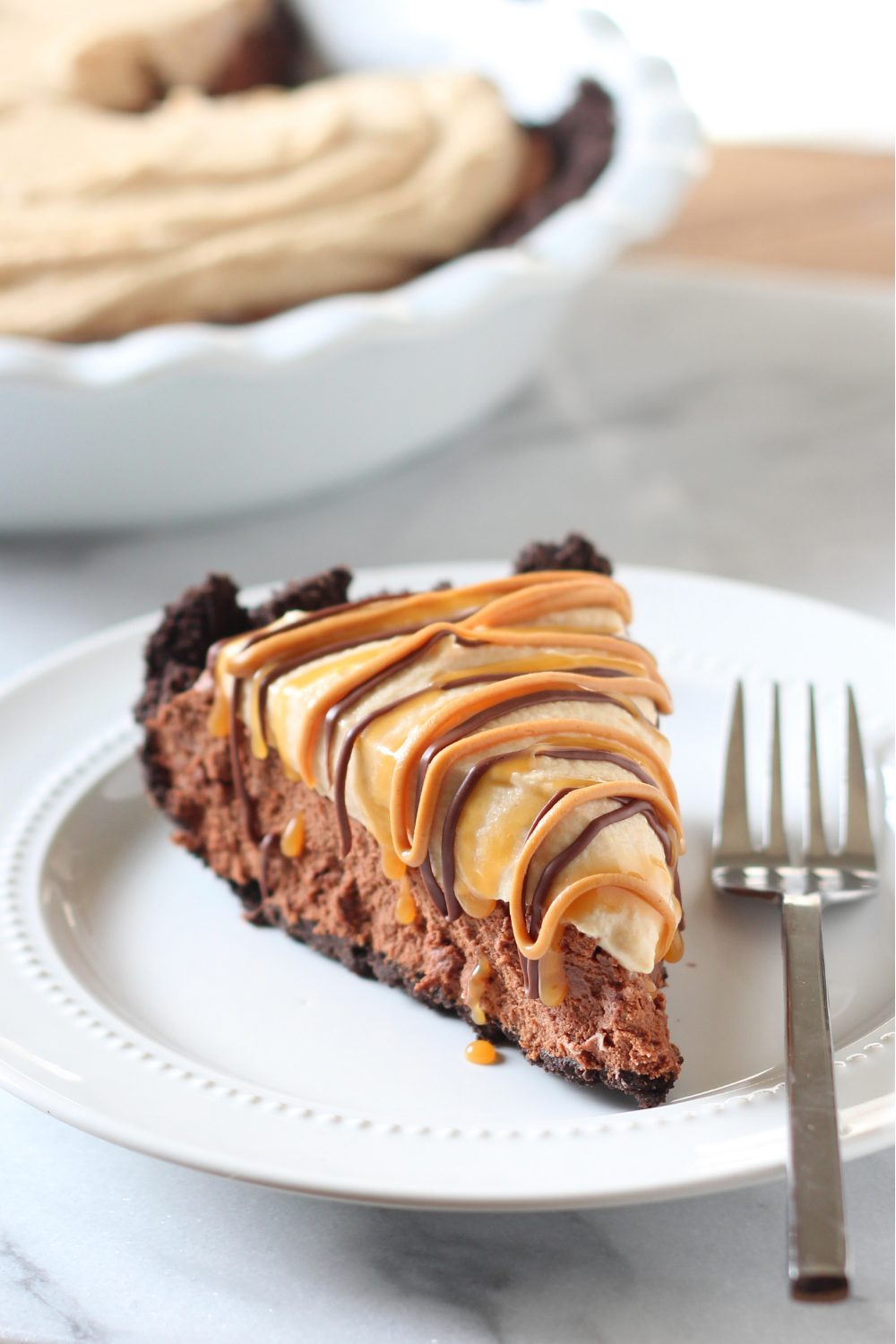 a closeup of Chocolate Peanut Butter Caramel Mousse Pie slice on a plate beside a fork, with the rest of the whole pie in the background