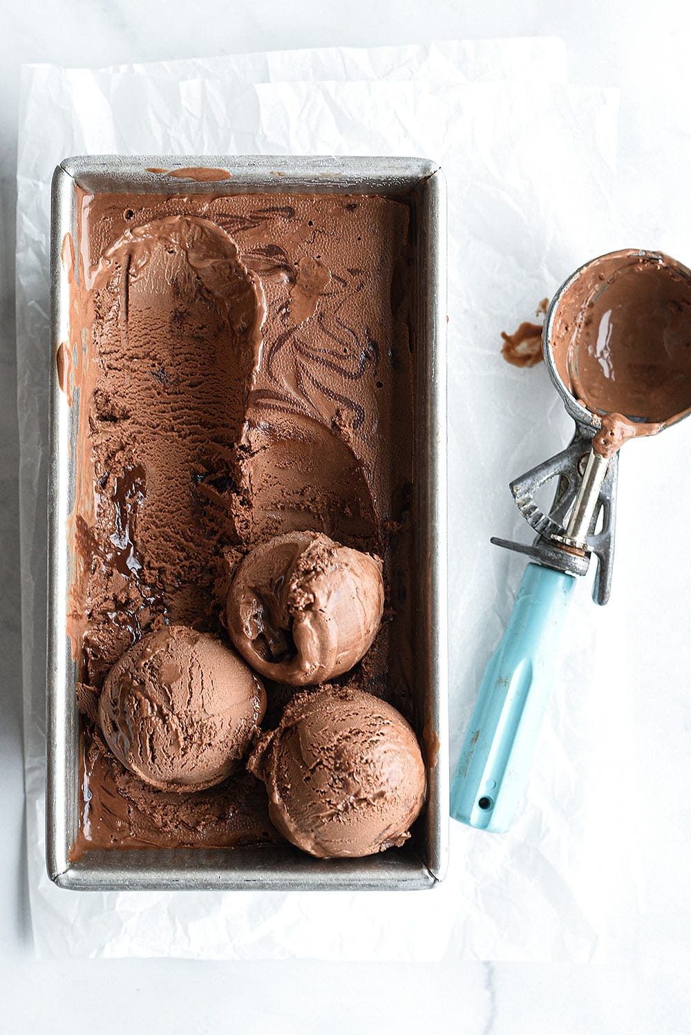 scoops of homemade chocolate ice cream in an ice cream tin with a blue ice cream scooper
