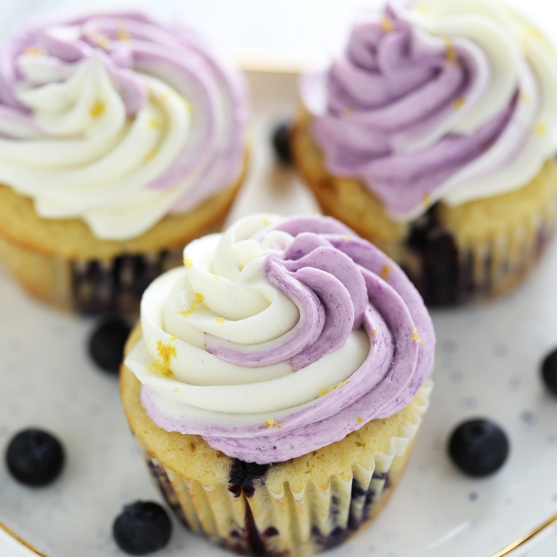three lemon blueberry cupcakes on a platter, with a few fresh blueberries scattered, ready to serve.