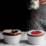 two Chocolate Soufflés in white ramekins, with powdered sugar being sprinkled across the tops.