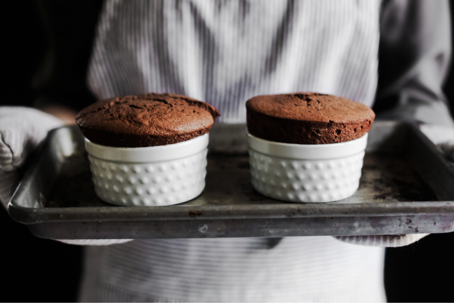 two Chocolate Soufflés in white ramekins on a baking tray, fresh out of the oven.