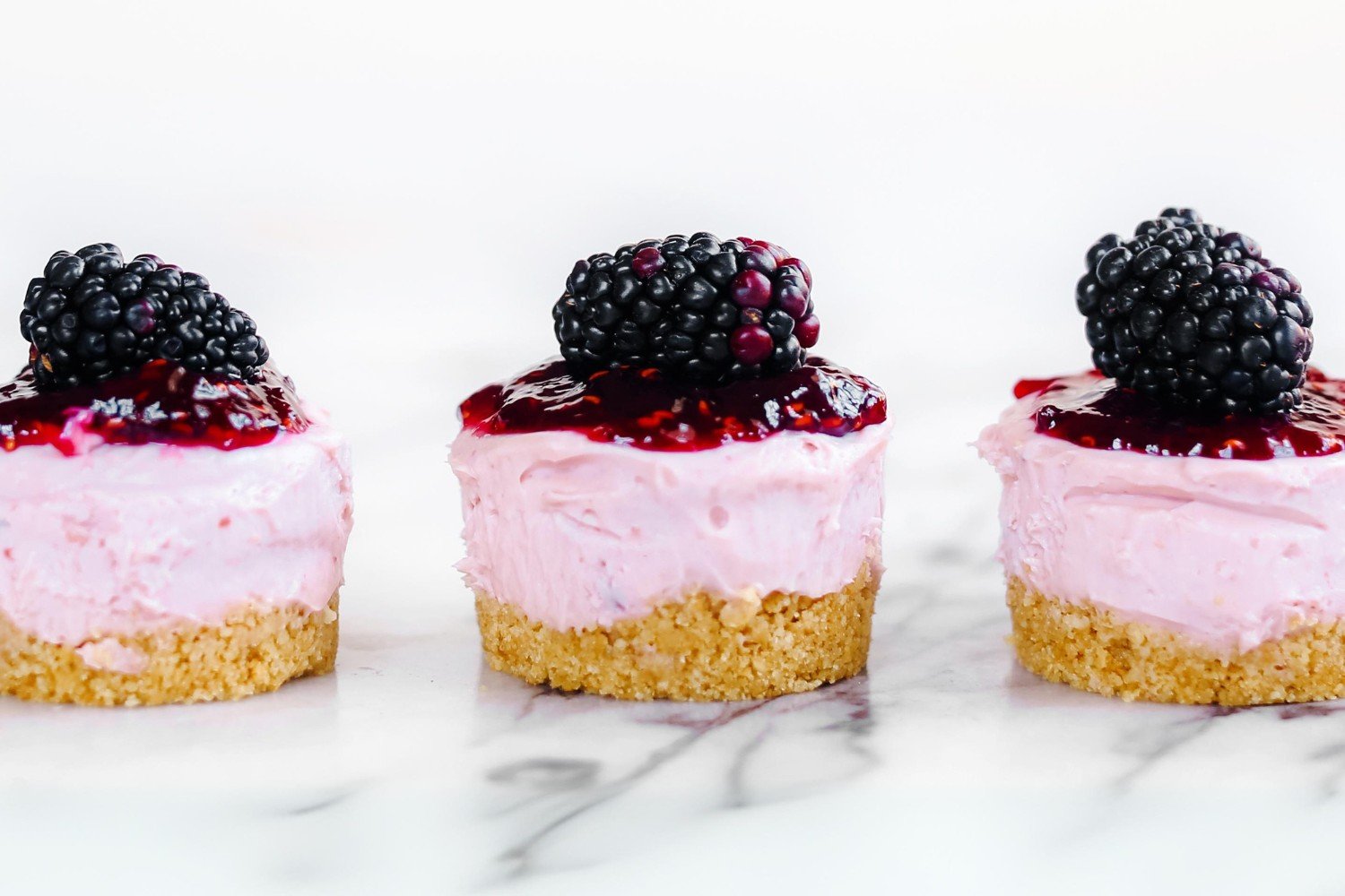 three three No Bake Mini Blackberry Cheesecakes in a row, with a fresh blackberry on top of each, ready to serve.