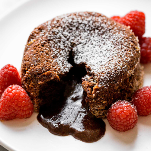 one chocolate lava cake, slightly broken open so the gooey interior is melting out, surrounded by a few fresh raspberries.