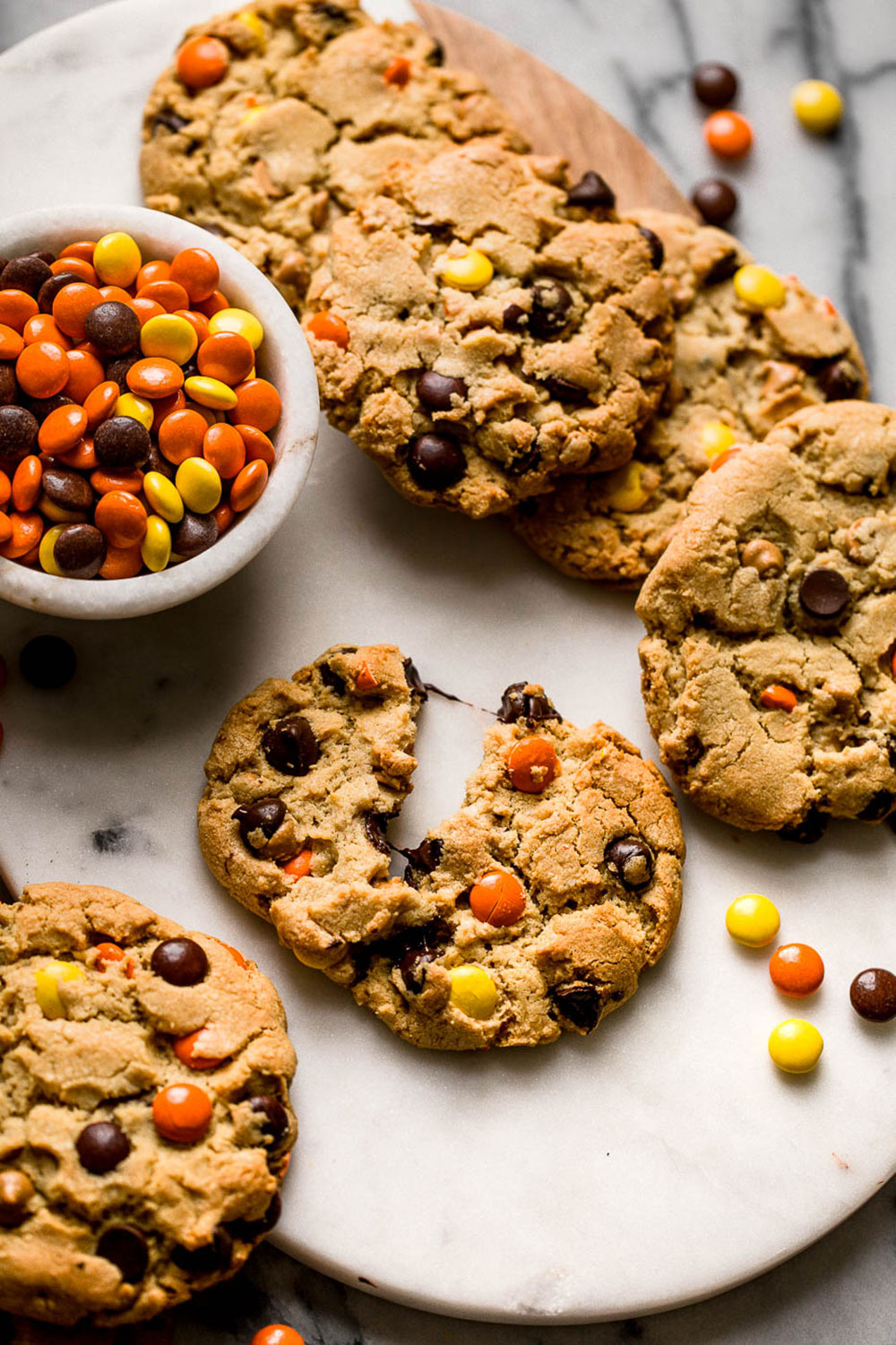 several Giant Reese's Pieces Chocolate Chip Cookies on a marble background, with one cookie broken in half and a bowl of Reese's Pieces to the side