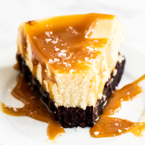 a slice of cheesecake with brownie base and topped with caramel sauce and flaked salt, sitting on a white plate, ready to serve.
