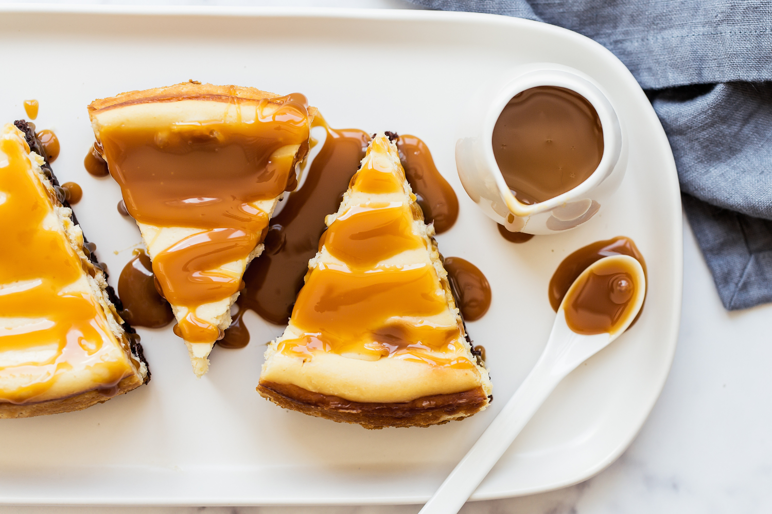 a few slices of Caramel Brownie Cheesecake on a white platter, with a small pot of caramel sauce to serve.