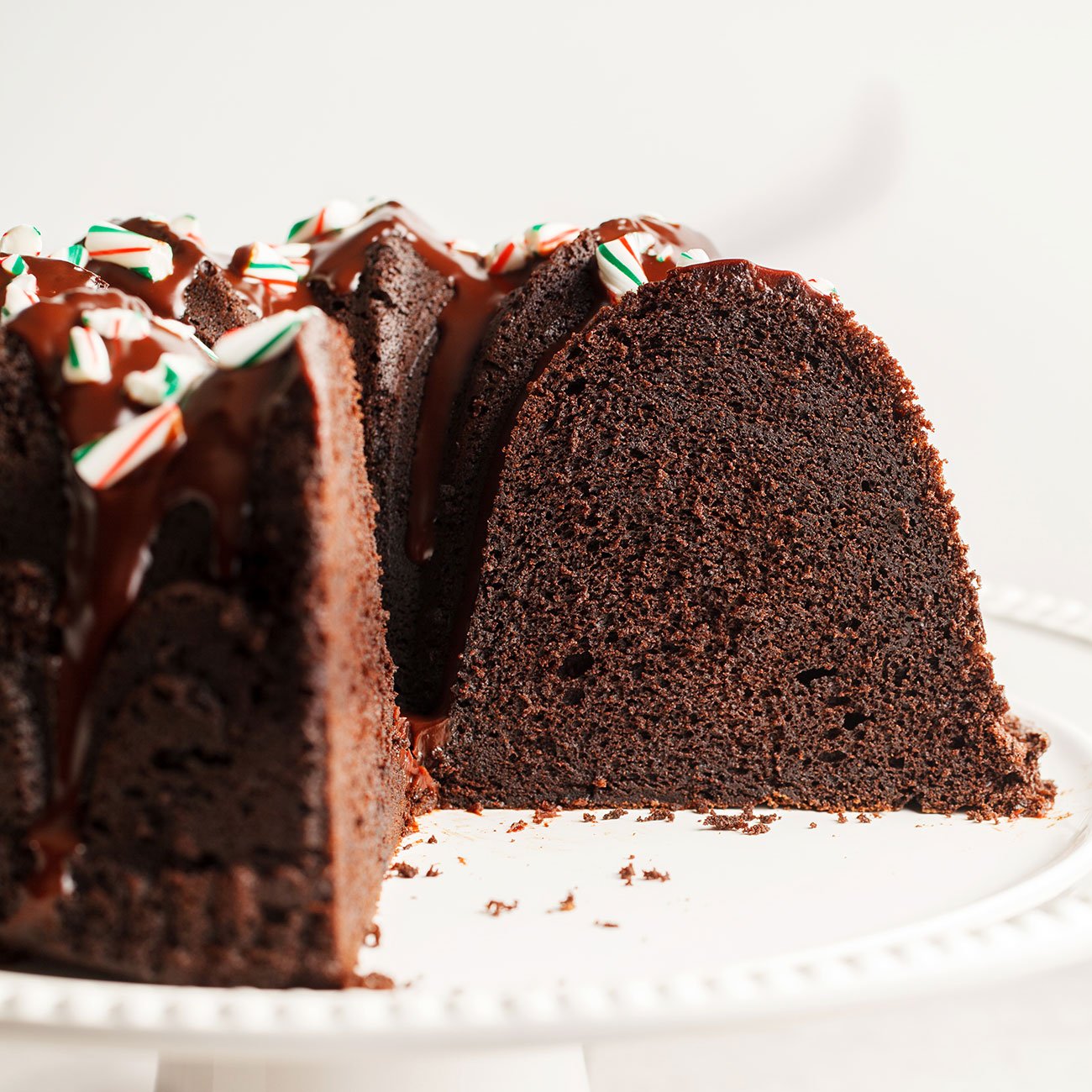 peppermint mocha bundt cake topped with chocolate ganache and crushed candy canes, placed on a white cake stand, with a slice taken out so you can see the moist interior.