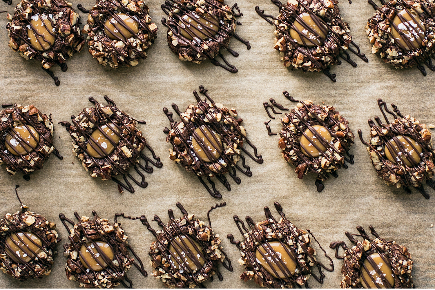straight-on top view of many Turtle Thumbprint Cookies on a parchment paper-lined baking tray, all drizzled with chocolate.