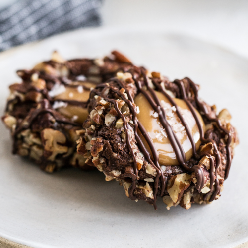 two thumbprint cookies on a white plate, rolled in nuts, filled with salted caramel, and drizzled with chocolate.