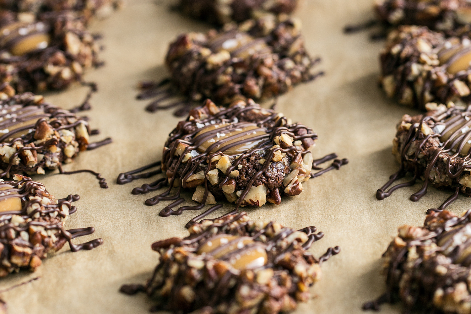 side view of many Turtle Thumbprint Cookies on a parchment paper-lined baking tray, all drizzled with chocolate.