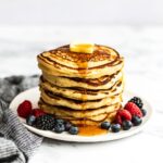 Tall stack of pancakes on a plate with syrup, berries, and a pat of butter