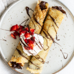 two crepes filled with Nutella, rolled and on a plate, drizzled with chocolate and topped with whipped cream and fresh strawberries.