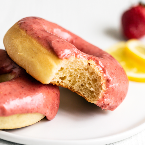 two strawberry lemon donuts on a white plate, one with a bite taken out.