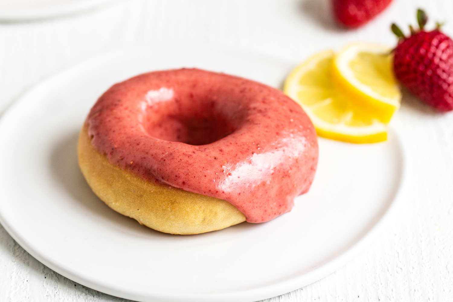 one Baked Strawberry Lemon Doughnut on a white plate with slices of lemon behind.