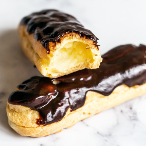 two eclairs on a marble surface, one with a bite taken out, showing the pastry cream inside.