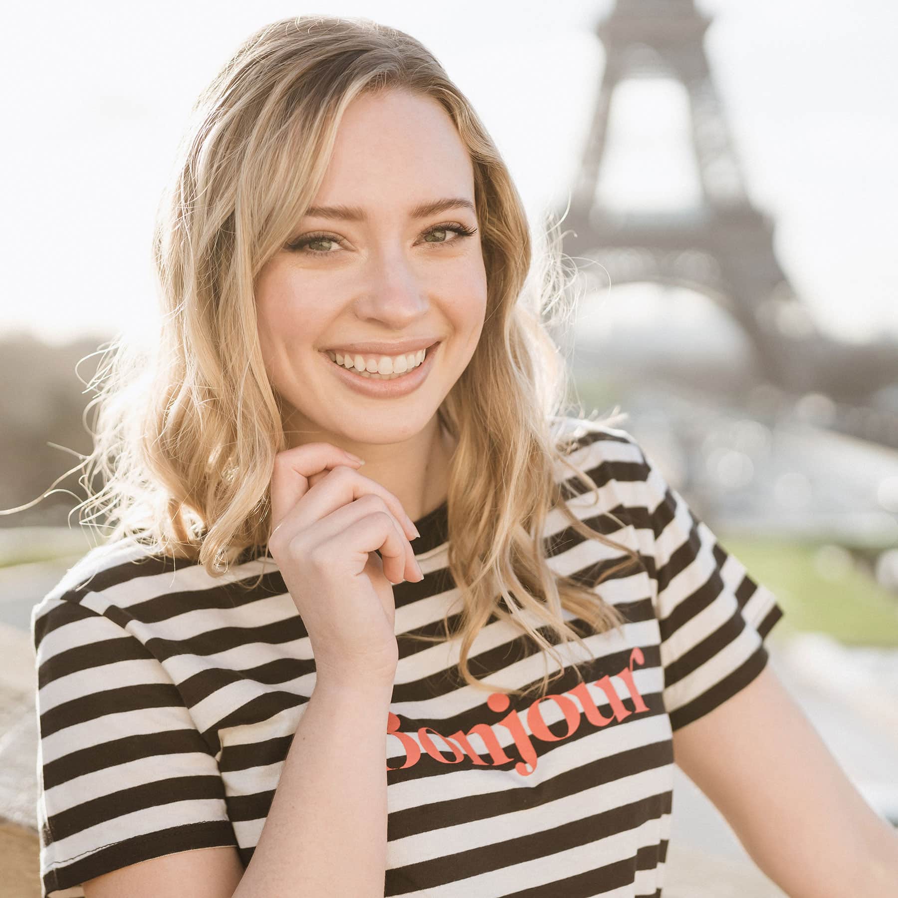 Tessa in front of the Eiffel Tower in Paris, France wearing a shirt that says Bonjour