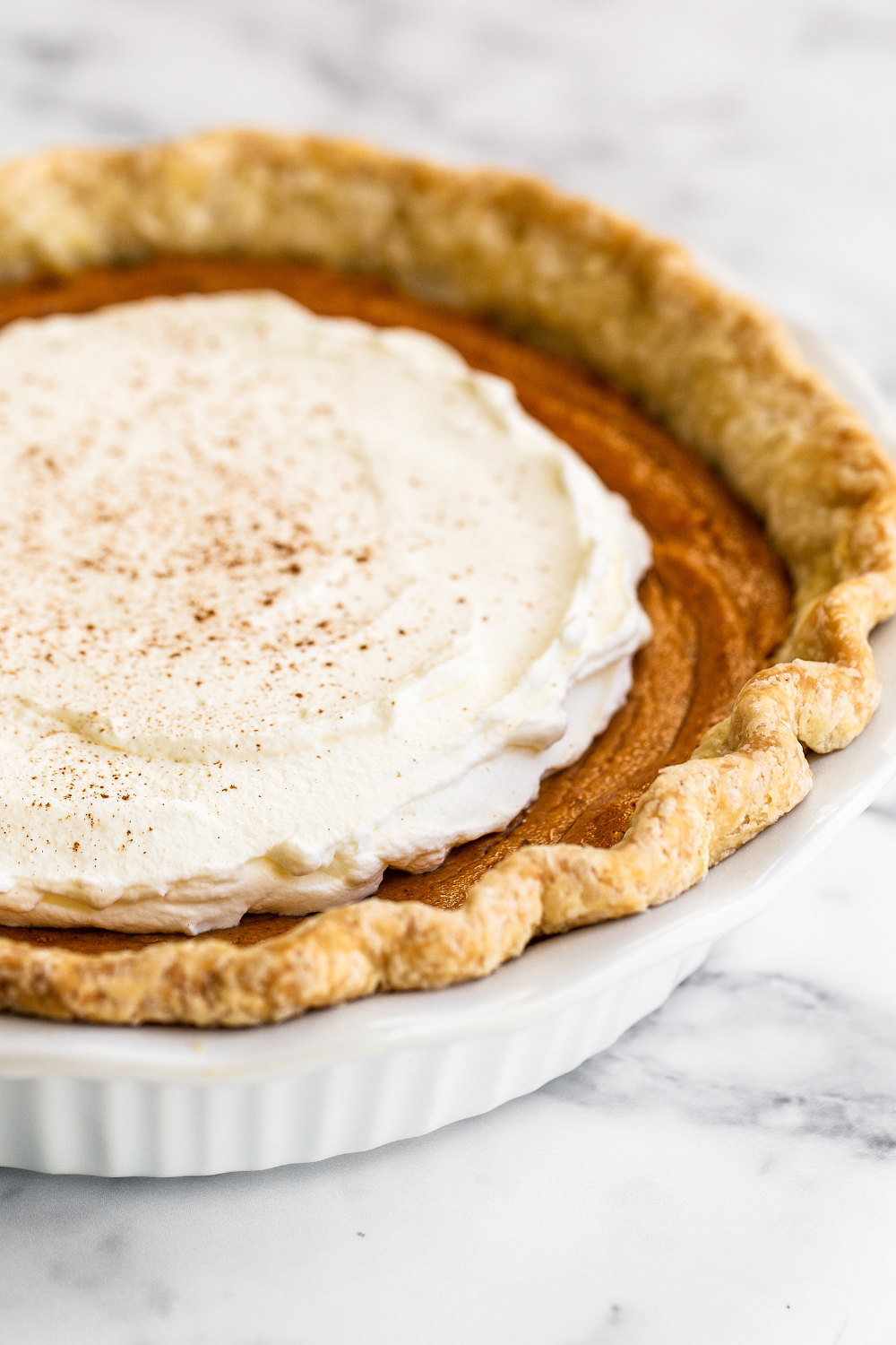 whole, unsliced Brown Butter Sweet Potato Pie in a white ceramic pie dish, with whipped cream and a hint of cinnamon sprinkled on top, ready to slice up and serve.
