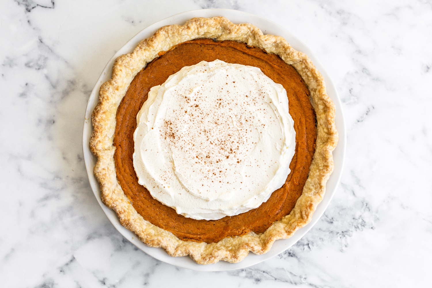 whole, unsliced Brown Butter Sweet Potato Pie in a white ceramic pie dish, with whipped cream and a hint of cinnamon sprinkled on top.