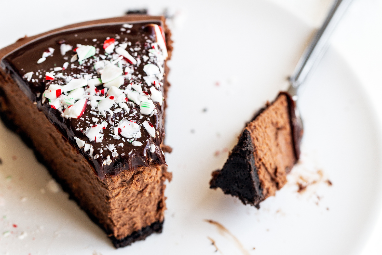 a slice of Chocolate Peppermint Cheesecake topped with ganache and crushed candy canes, on a plate with a fork, ready to take a bite.