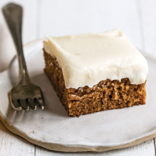 slice of gingerbread sheet cake with cream cheese frosting on a plate, with a fork beside.
