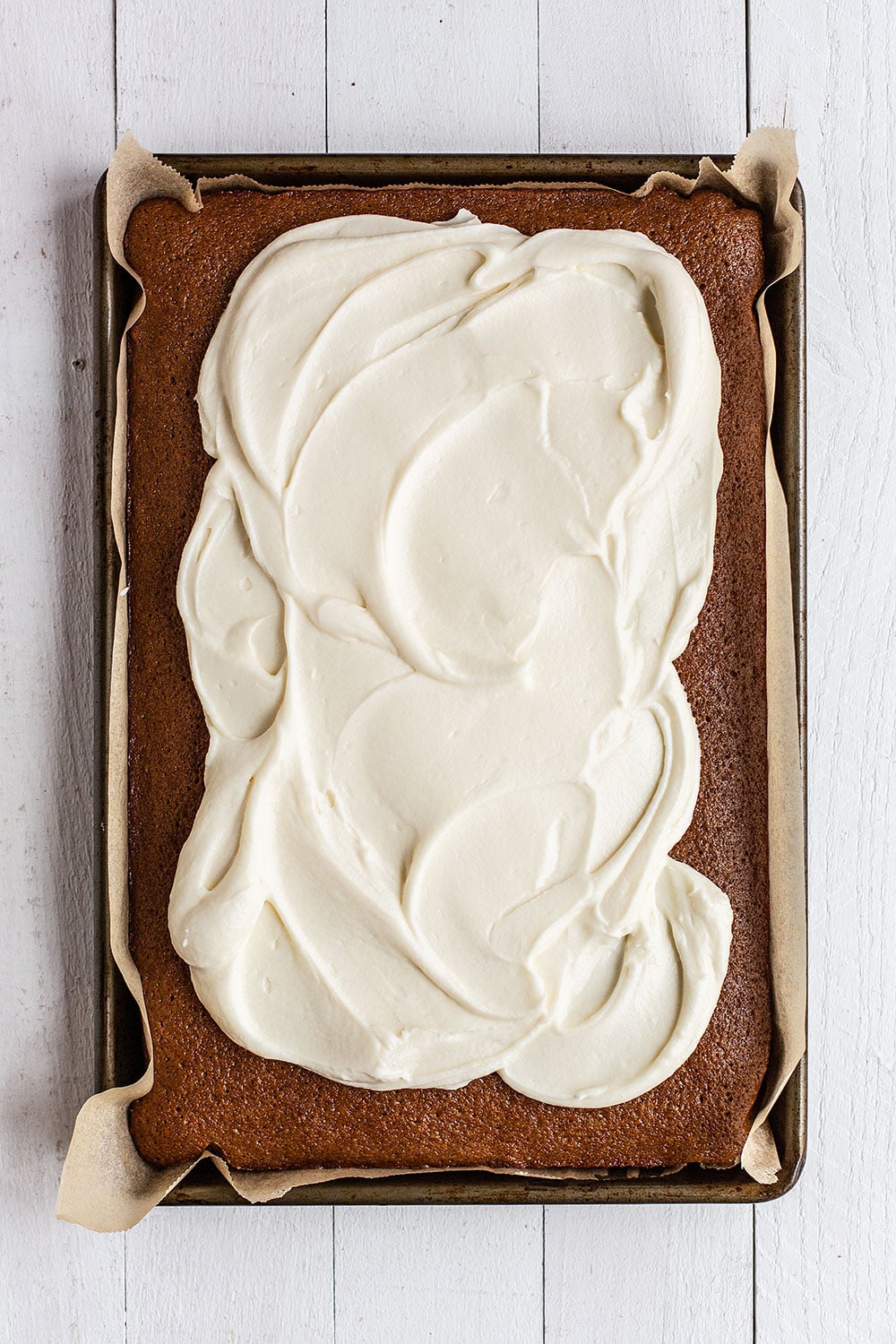 Gingerbread sheet cake in baking pan with cream cheese icing being spread on top