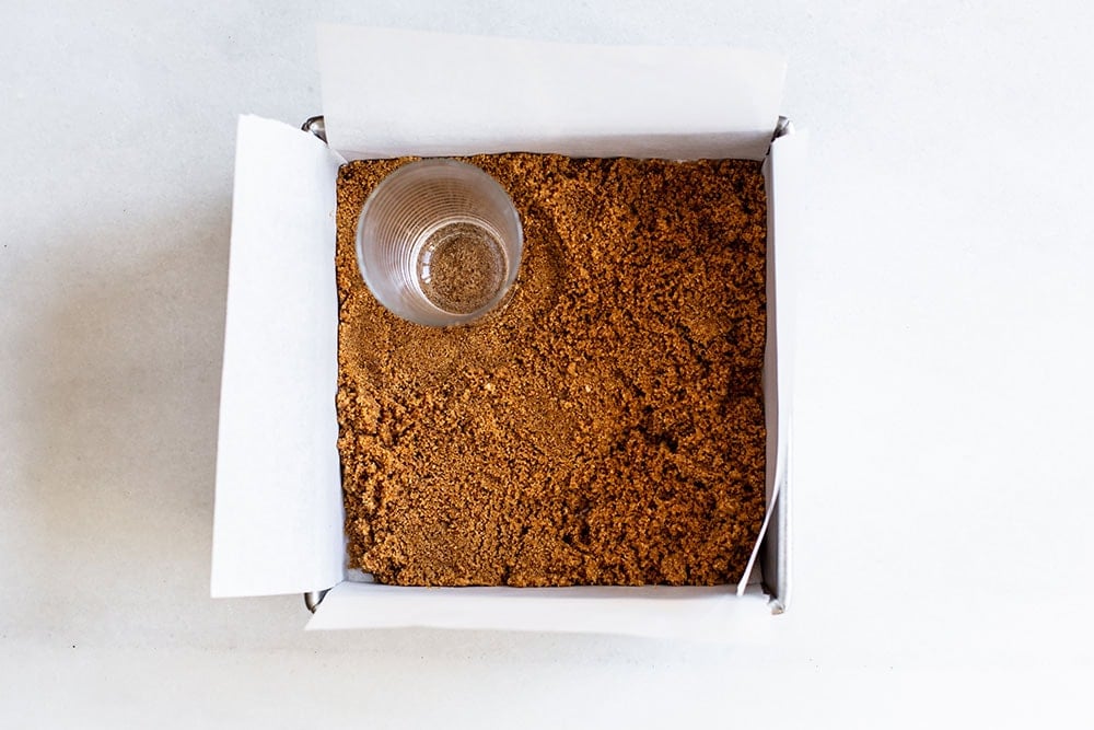 Graham cracker crust being pressed into a baking pan with a cup