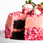 rich chocolate cake with strawberry buttercream between the layers and frosting the whole cake, with Valentine's sprinkles across the bottom of the cake, and chocolate-dipped strawberries on top, with a slice taken out so you can see the cake inside.