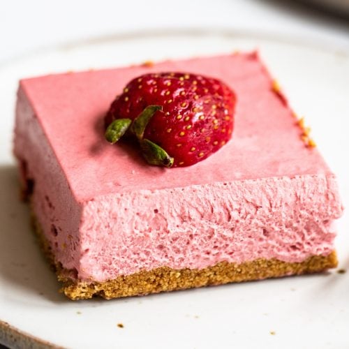a square of strawberry cheesecake bar on a white plate with half a fresh strawberry on top.