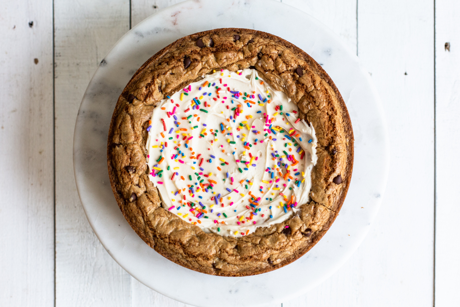 a round chocolate chip cookie cake with vanilla buttercream frosting and sprinkles on top, sitting on a marble cake stand
