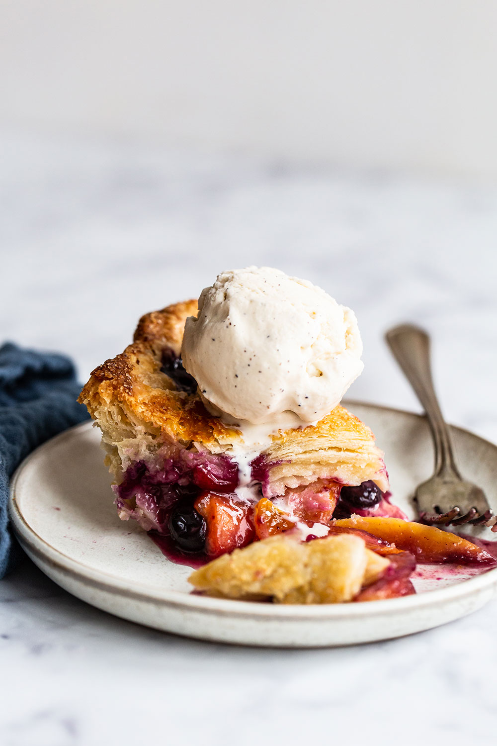 slice of blueberry peach pie with flaky crust, topped with a scoop of vanilla ice cream