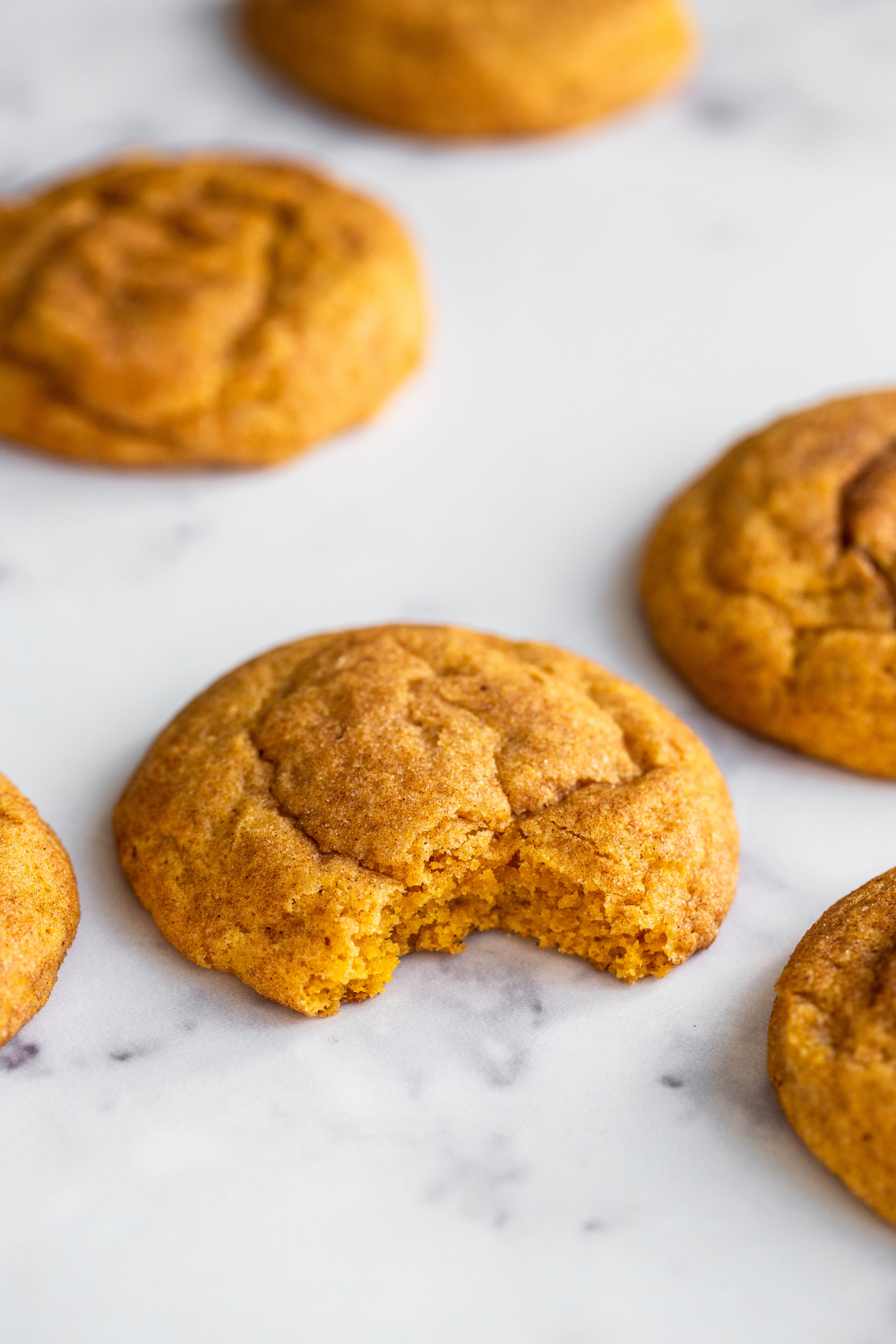 several pumpkin snickerdoodle cookies on a marble background, with one cookie with a bite taken out