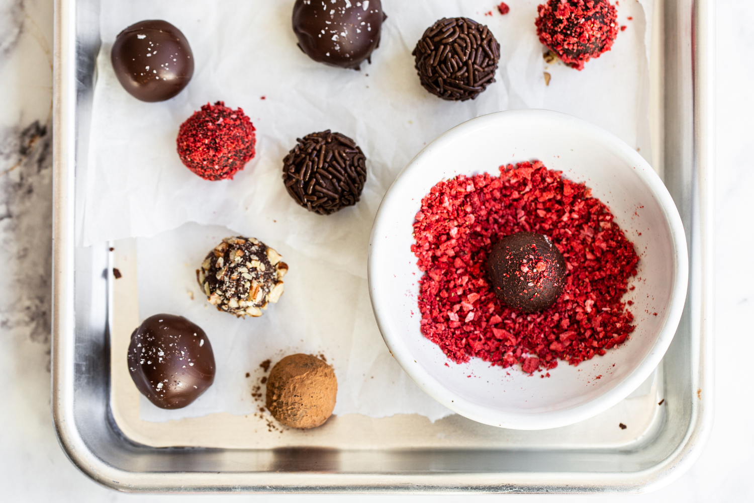 a tray of truffles coated in nuts or chocolate sprinkles, with one truffle being coated in crushed freeze-dried raspberries.