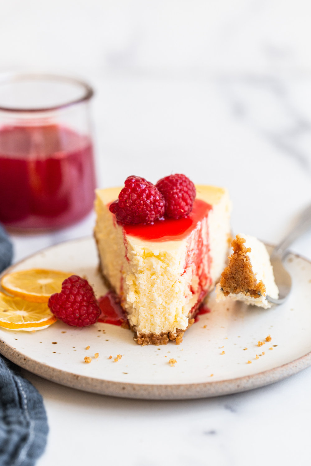 slice of cheesecake with a bite taken out, topped with raspberry sauce and fresh raspberries, on a plate, ready to be enjoyed