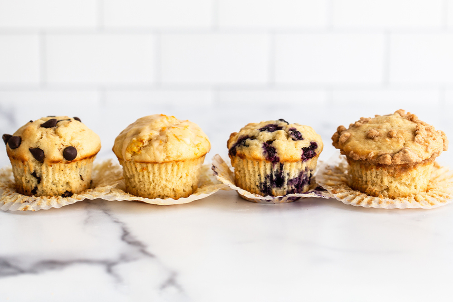 lineup of tall bakery style muffins in muffin liners on a white marble surface.