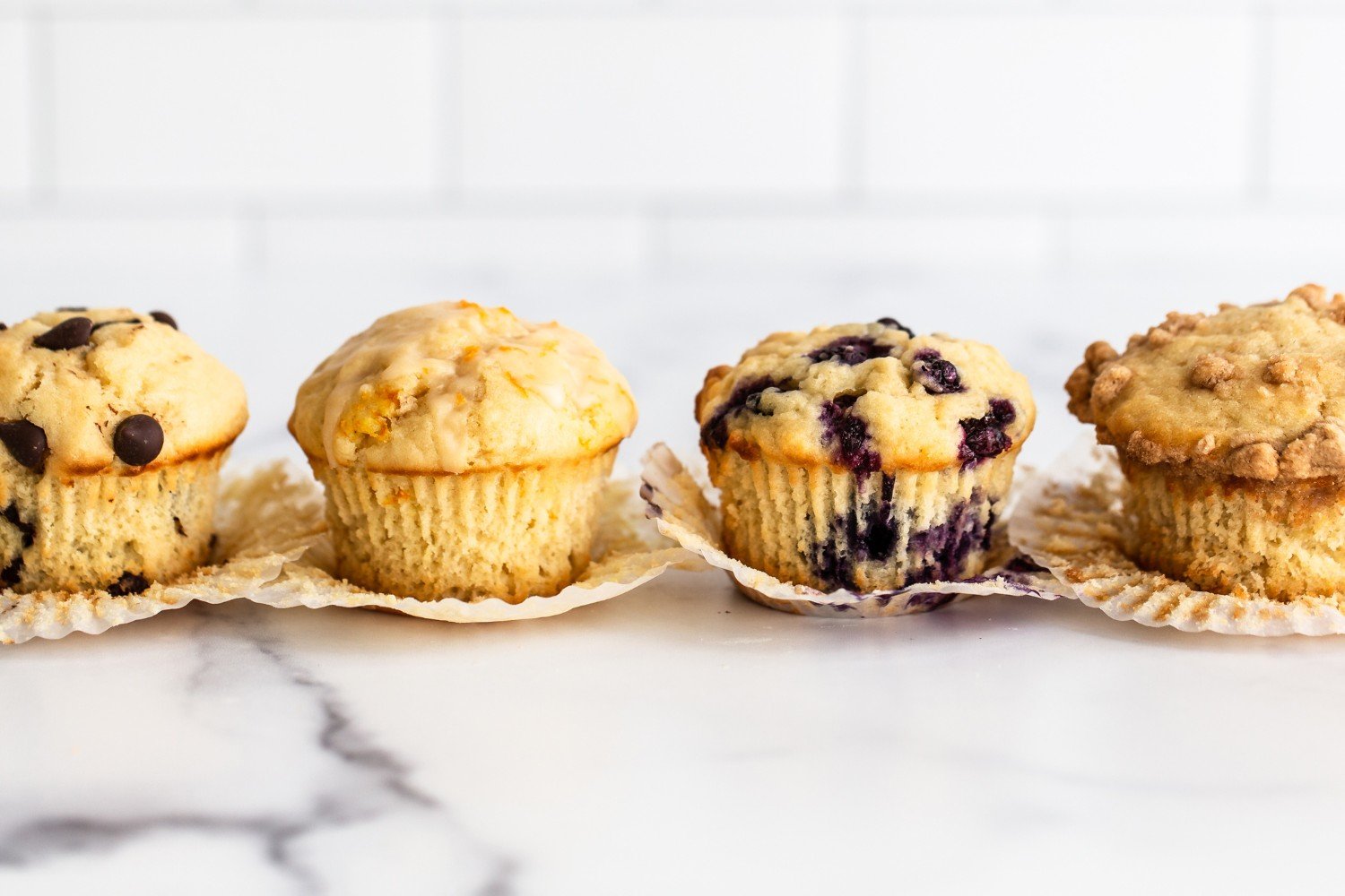 a lineup of four different types of muffins in a row on a white tile background.