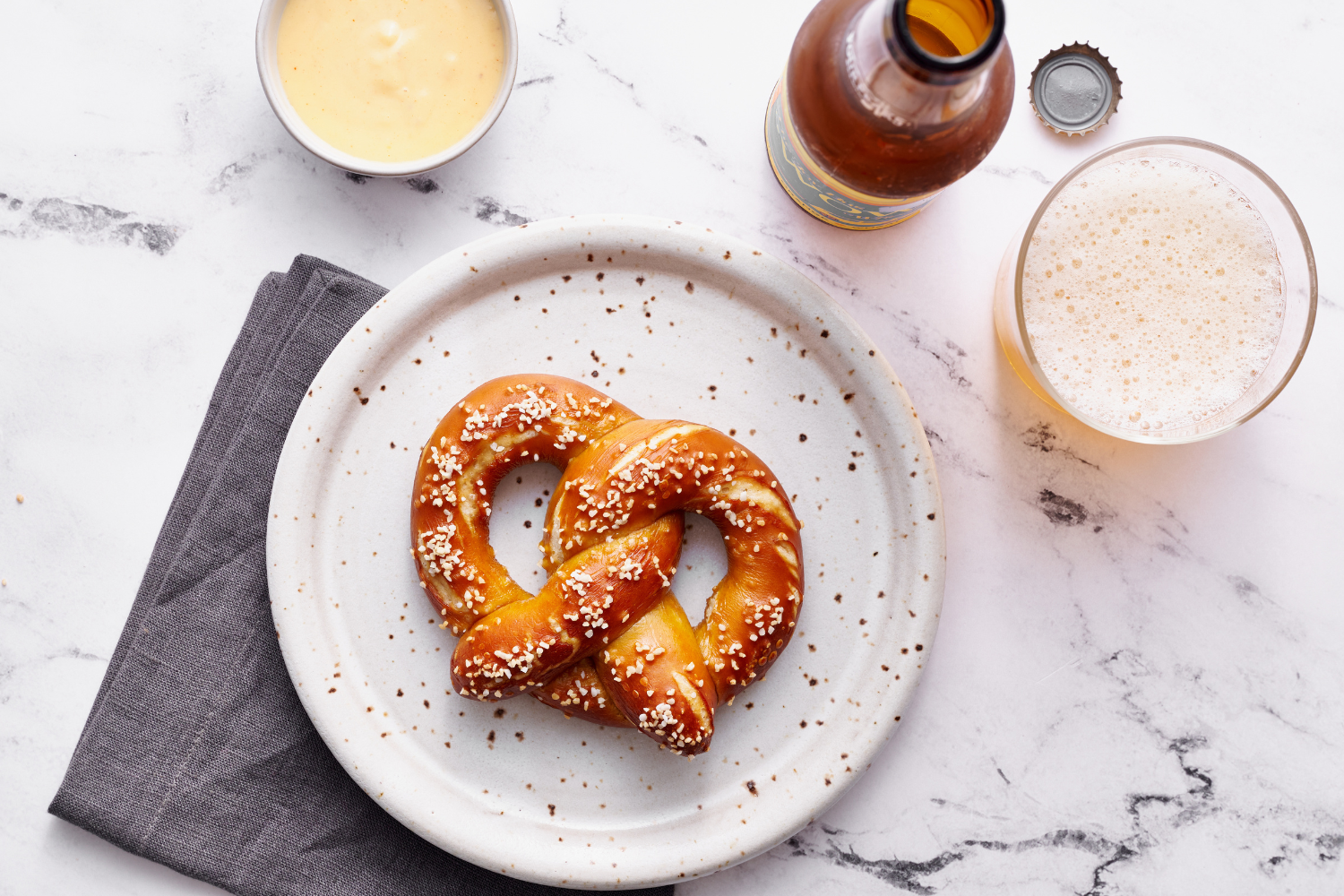 a fresh soft pretzel with coarse salt on a plate, with a side of beer cheese and a bottle of beer
