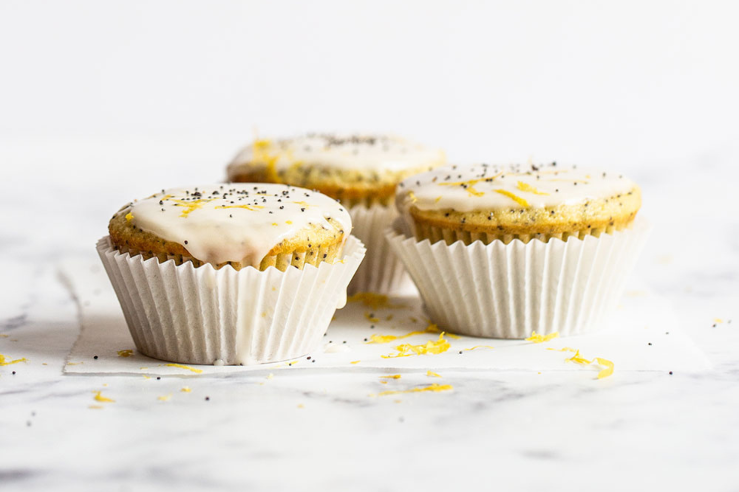 three glazed lemon poppyseed muffins in muffin liners, sitting on a marble counter