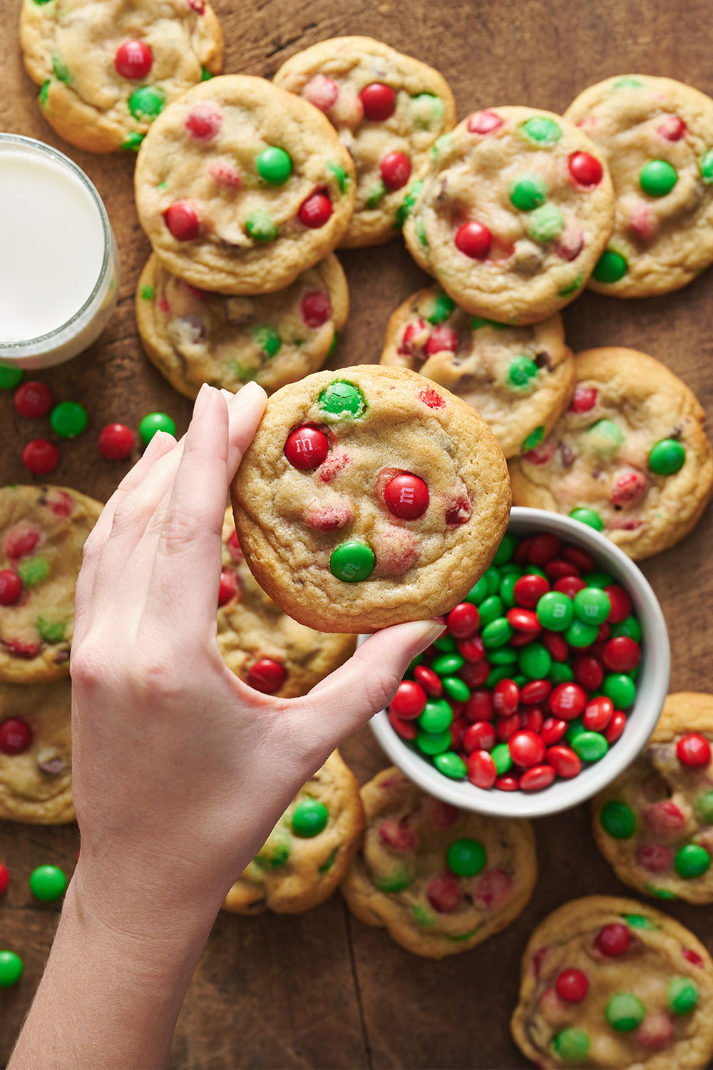 an array of M&M Christmas Cookies with a bowl of red and green M&Ms, and a hand holding one of the cookies in the foreground.