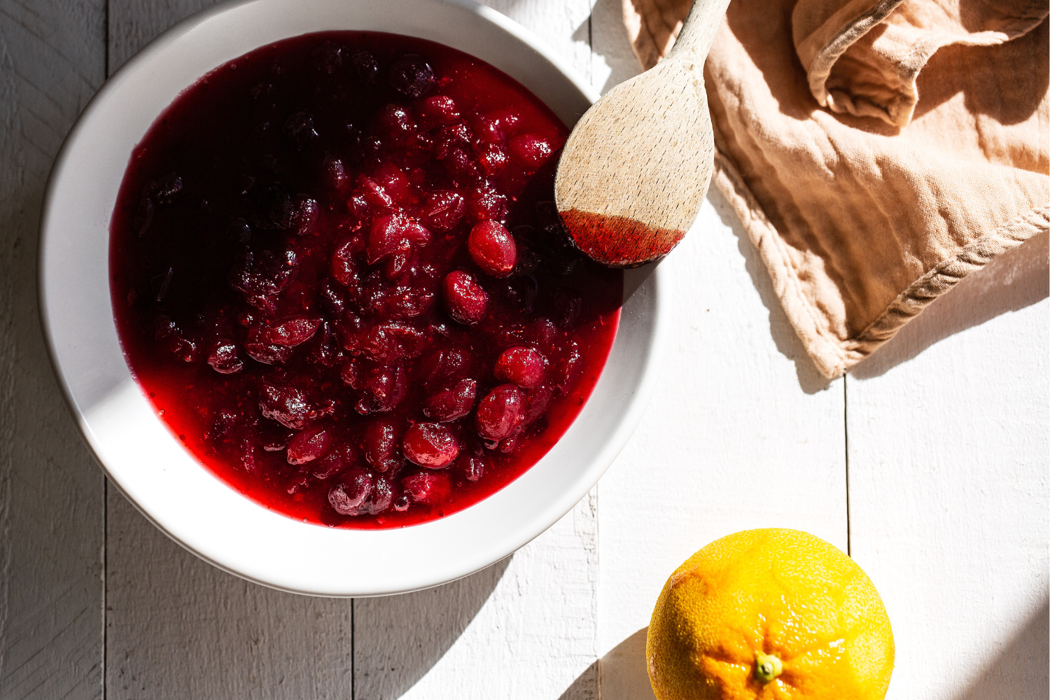 a bowl of cranberry sauce with a wooden spoon, next to an orange and a napkin.