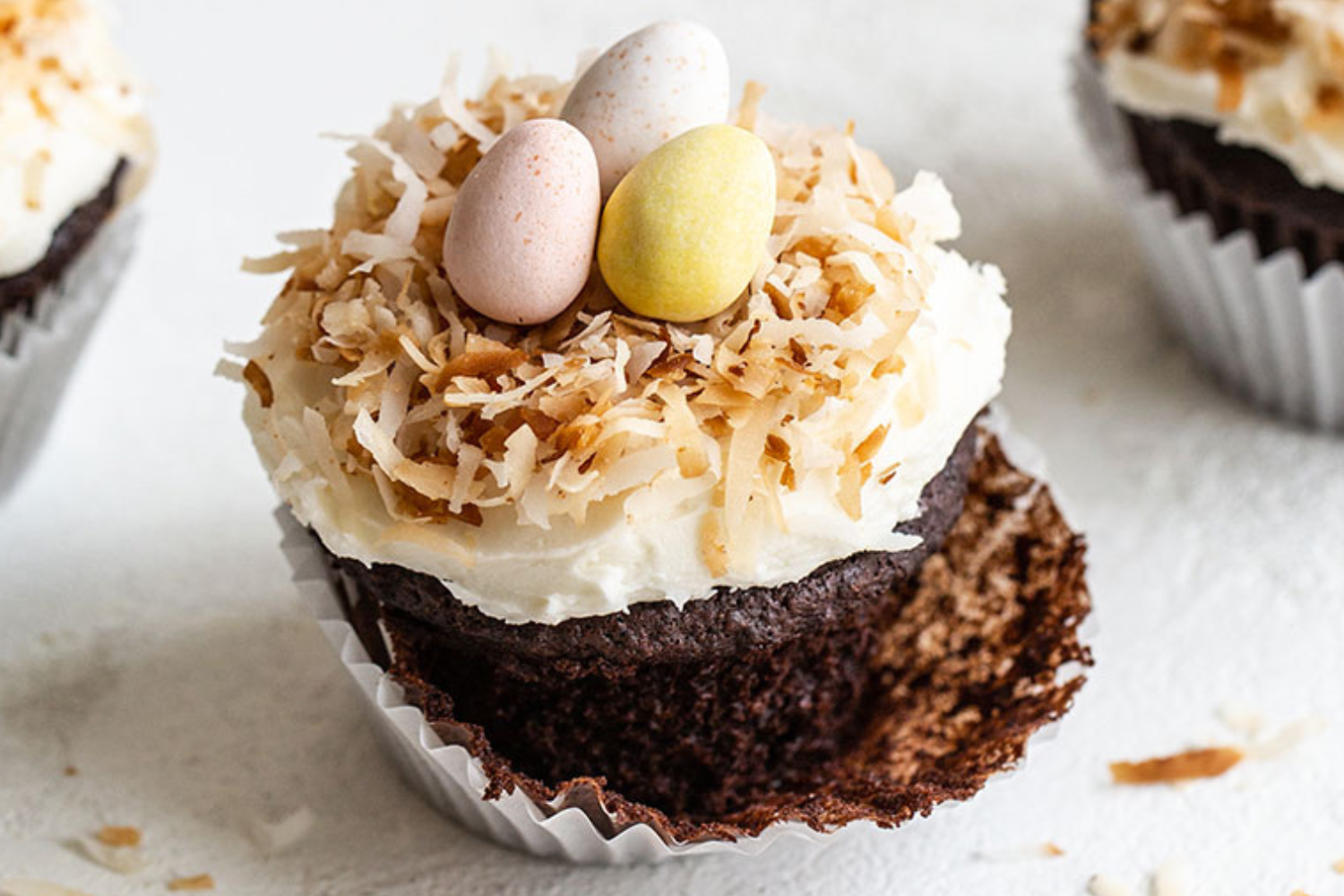 a chocolate cupcake with coconut frosting, topped with toasted coconut and chocolate eggs, with its wrapper half removed, ready to enjoy.