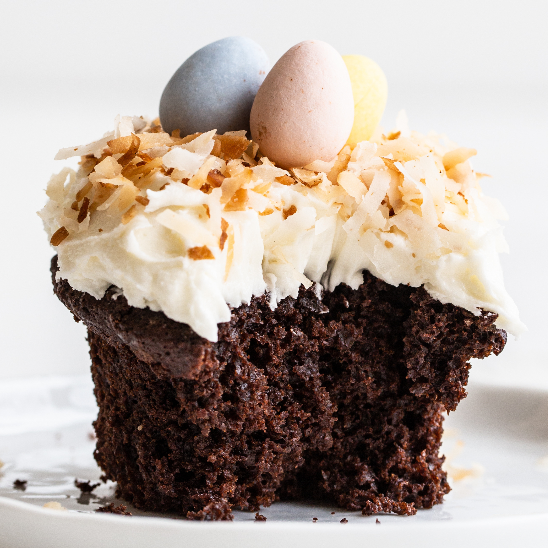 a chocolate cupcake with coconut frosting, topped with toasted coconut and chocolate eggs, with a bite taken out.