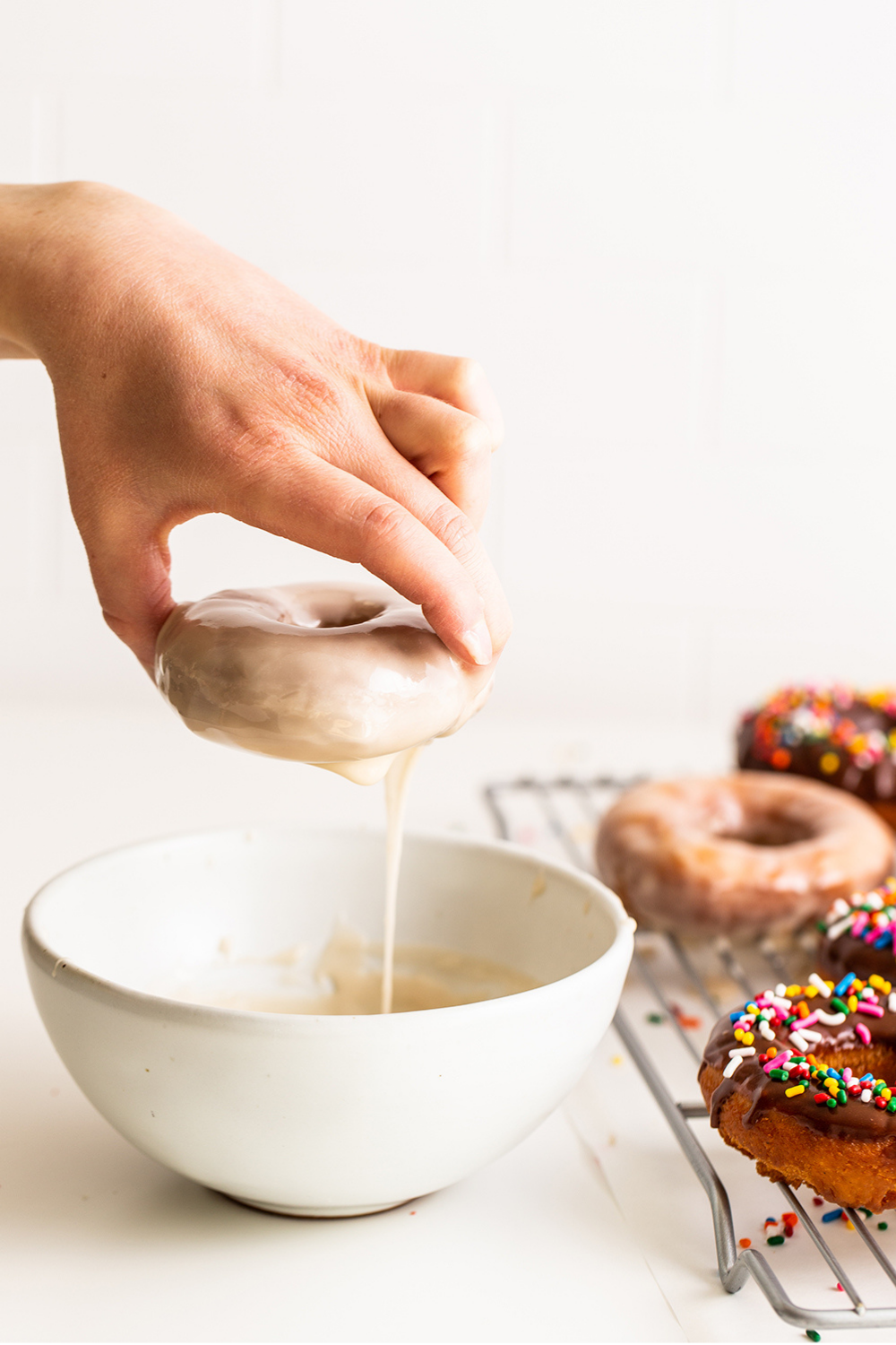 a hand dipping a donut into a bowl of glaze and allowing the excess to drip back into the bowl.