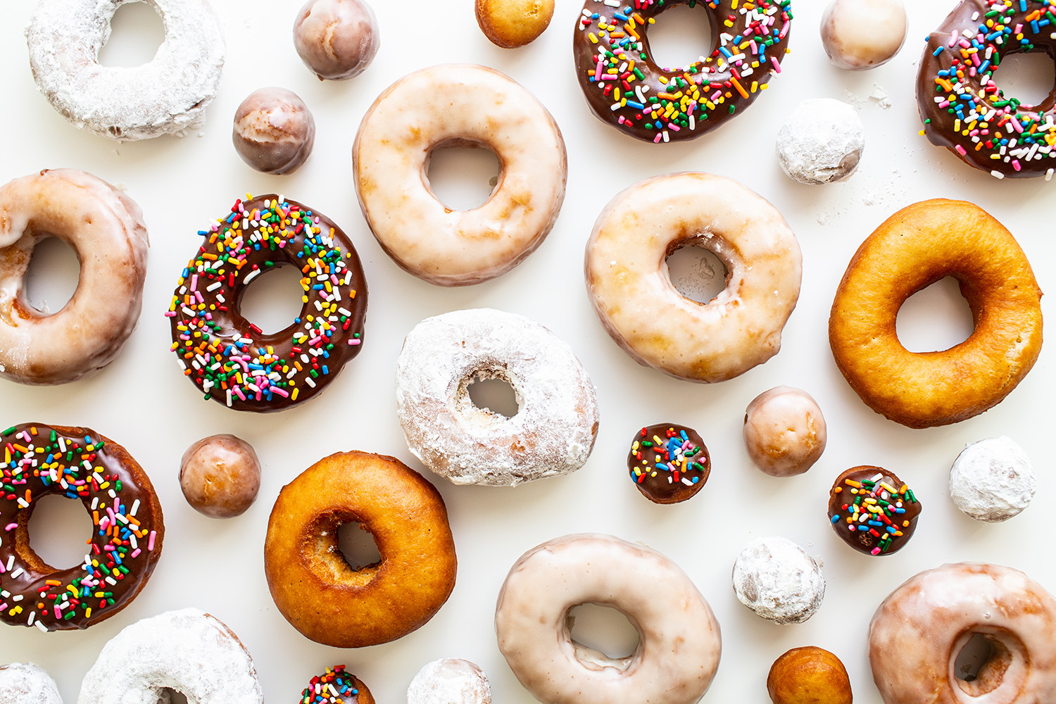 a dozen doughnuts on a white background, with some doughnut holes scattered between.