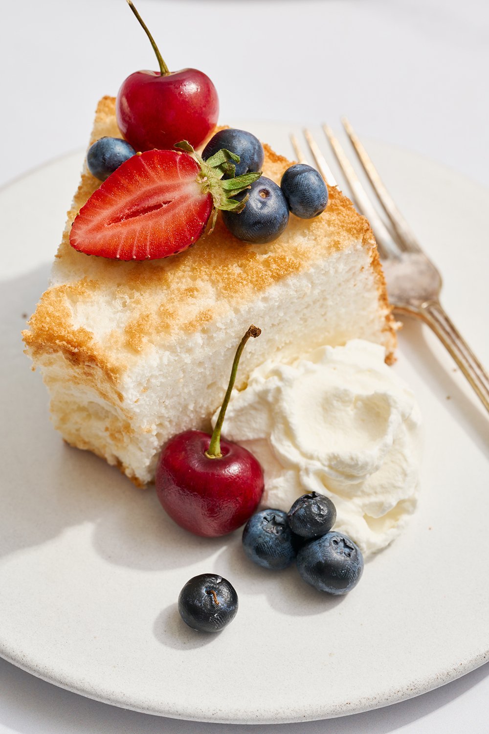 slice of angel food cake on a white plate with a fork, and whipped cream and berries to serve
